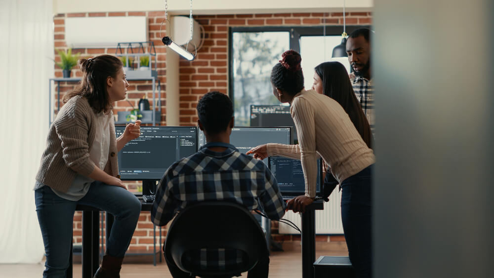 Equipe de desenvolvedores colabora em frente a computadores com códigos na tela, representando o trabalho em uma software house.